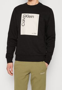 Felpa nera con girocollo e maniche lunghe. Presenta una grande grafica quadrata bianca con il testo "Calvin Klein" e "Est. 1968".