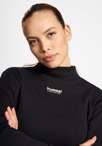 Zwarte geribbelde langemouwen top met hoge kraag, voorzien van een witte "hummel SPORTSWEAR" logo op de borst. Soepele, zachte textuur.