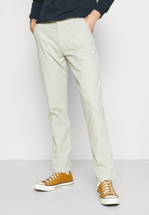Man die beige broek met rechte pijpen draagt, handen in zijn zakken en mosterdgele hoge sneakers, staand tegen een witte achtergrond.