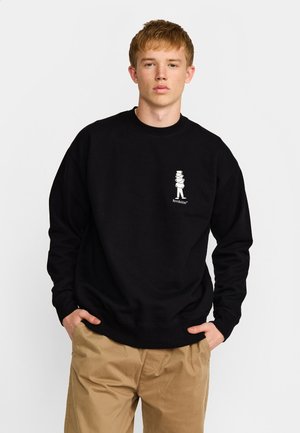 Jonge man met kort rood haar, draagt een zwarte trui met een kleine witte afbeelding en beige broek, staand met handen in de zakken.