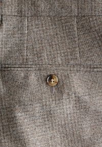 Brauner, strukturierter Stoff mit einem Fischgrätenmuster, ausgestattet mit einem großen, glänzenden Knopf, der auf die Vorderseite genäht ist, sowie sichtbaren Nahtdetails.