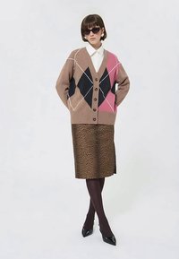 Moteris vilki didelį rudą kardiganą su rožiniais ir juodais deimanto raštais, baltą marškinėlį, leopardų rašto sijoną, juodas pėdkelnes ir aukštakulnius batus.