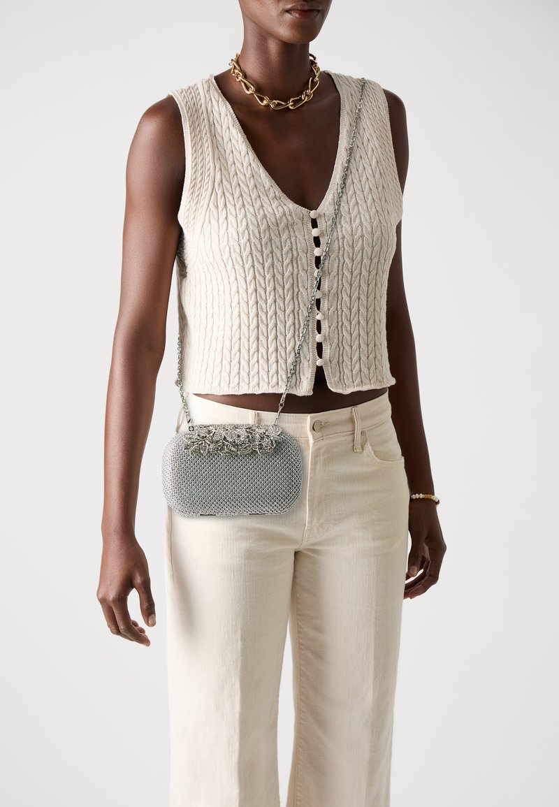 Bolso de mano metálico plateado con textura de malla, adornado con flores en la parte superior, combinado con un chaleco de punto crema y pantalones de color claro.