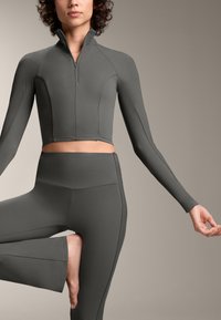 Sivá atletická krátka bunda s polovičným zipsom a rebrovanou textúrou, v kombinácii s legínami s vysokým pásom, ktoré majú rozšírené konce a bočné detaily.