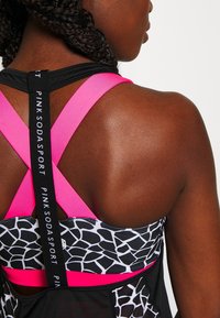 Sportovní podprsenka s černým základem, růžovými popruhy a vzorovaným černobílým designem. Obsahuje styl s křížovým záznamem a logo na popruzích.