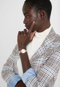 Goldene Armbanduhr mit rundem Ziffernblatt, getragen auf einem dunklen Hautton. Der Träger trägt einen hellen Rollkragenpullover und ein kariertes Blazer mit hellblauen Akzenten.