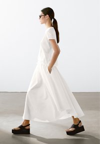 Hvid ankel-længde nederdel med draperet design, parret med en tætsiddende hvid t-shirt og sorte platform sandaler. Glatte, minimalistiske teksturer.