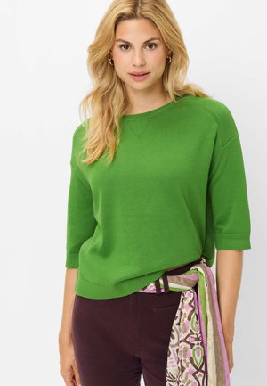 Lyshåret kvinde iført en grøn kortærmet sweater, mørke bukser og et mønstret tørklæde bundet om taljen, stående foran en ensfarvet baggrund.