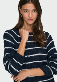 Pull marin avec des rayures horizontales blanches, en tissu tricoté texturé. Dispose d'un col rond et de manches longues avec des poignets côtelés.