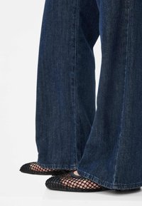 Nohy obuty v černých síťovaných balerínách v kombinaci s širokými tmavomodrými džíny na bílém pozadí.