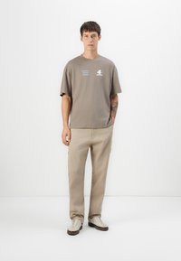 Gray cotton t-shirt with short sleeves, featuring blue and white text. Beige pants with side pockets, worn with cream shoes and brown accents.