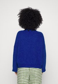 Maglione blu, strutturato e di grandi dimensioni, realizzato in maglia, con scollatura rotonda e spalle scese, indossato sopra pantaloni a quadri verde e bianco.