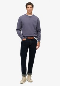 Bluza w stonowanym fioletowym kolorze z okrągłym dekoltem i wypukłym logo; zestawiona z ciemnymi dżinsami i beżowymi wysokimi trampkami. Brązowy skórzany pasek.