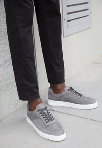 Szare zamszowe trampki z białymi gumowymi podeszwami, okrągłym noskiem i czarnymi sznurowadłami. Design cechuje się gładką teksturą i minimalnymi detalami.