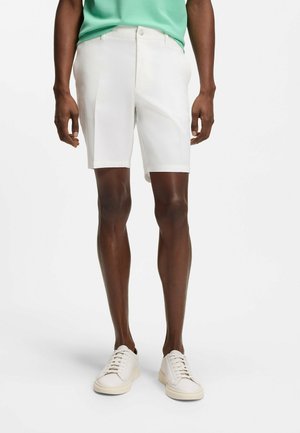 Mann trägt weiße, maßgeschneiderte Shorts, weiße Sneaker und ein grünes Hemd, steht vor einem einfachen weißen Hintergrund.