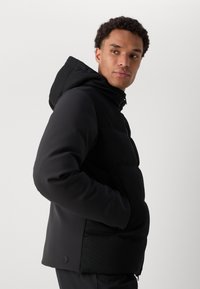 Black puffer jacket with a textured fabric panel, featuring a hood, zip closure, and side pockets. Smooth finish with subtle branding detail.
