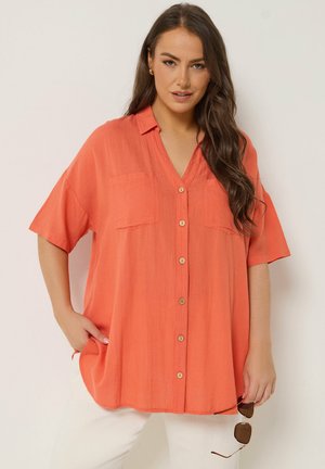 Vrouw met lang bruin haar draagt een losse oranje blouse met knopen en witte broek, houdt een zonnebril in haar rechterhand, staat voor een effen achtergrond.