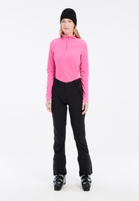 Pullover in pile di un brillante rosa con colletto a mezza zip, abbinato a pantaloni da sci neri con tasche con zip e vita aderente.