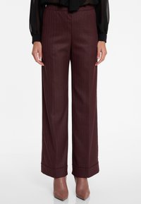 Vrouw in bordeauxrode wijde pijpen met streepdessin en met omslag afgewerkte zoom, met taupe puntige enkellaarzen, op een witte achtergrond.