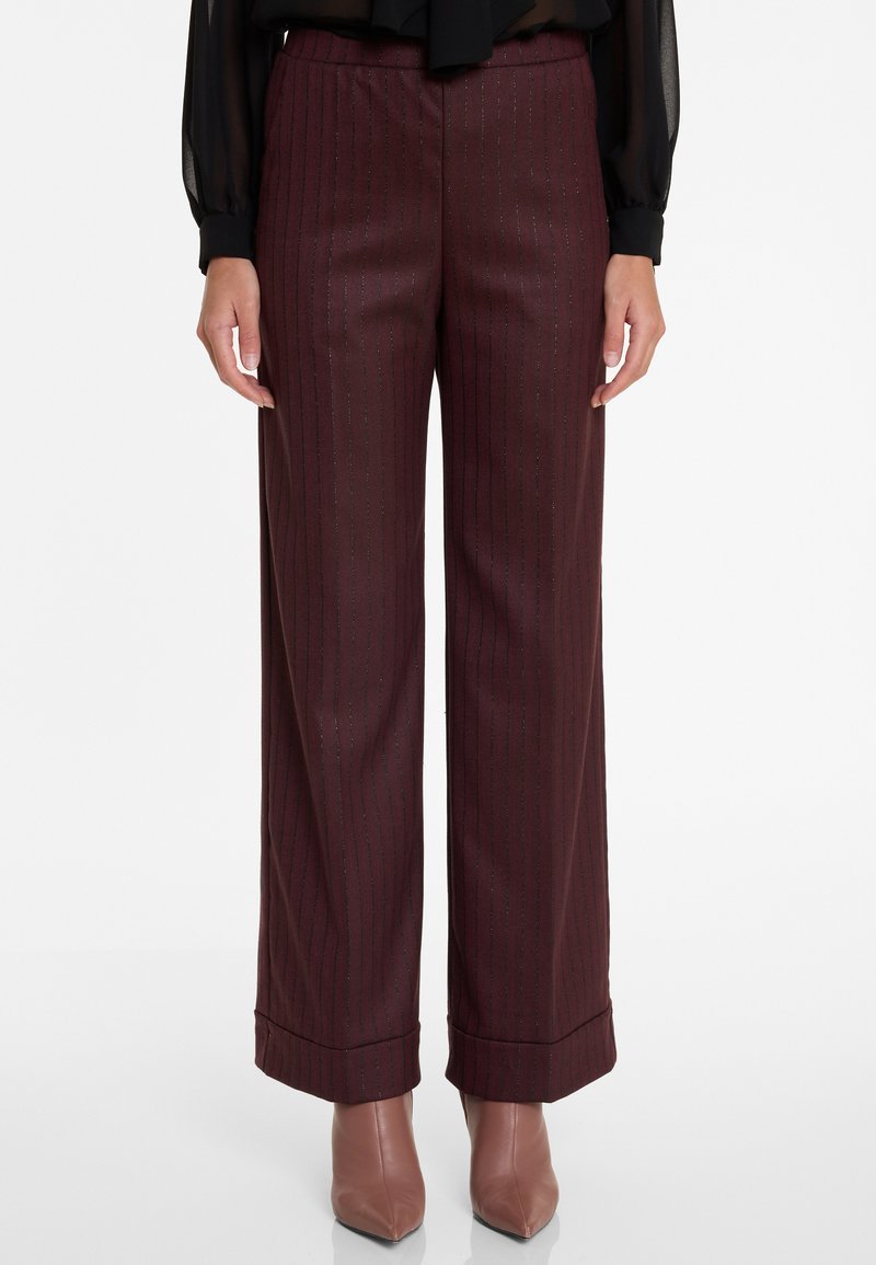 Vrouw in bordeauxrode wijde pijpen met streepdessin en met omslag afgewerkte zoom, met taupe puntige enkellaarzen, op een witte achtergrond.