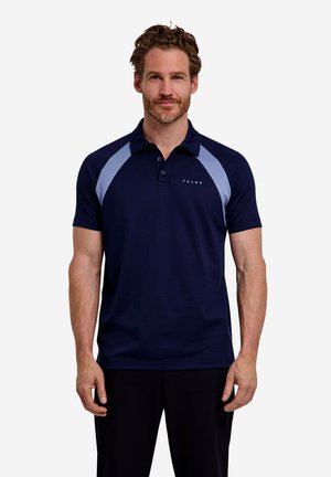 Mann mit lockigem Haar und Bart, der ein dunkelblaues Polohemd mit hellblauen Schulterpaneelen und schwarze Hose trägt, vor einfarbigem Hintergrund steht.