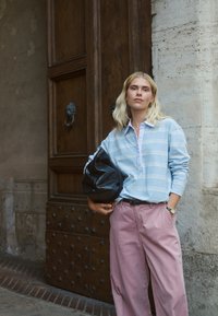 Mladá žena s blond vlasmi stojí pri veľkých drevených dverách, oblečená v modrej pruhovanej košeli a ružových nohaviciach, drží veľkú čiernu kabelku.