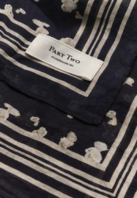 Mørk navy tørklæde med et abstrakt creme mønster, samt et fremtrædende mærke med teksten "PART TWO" syet på et lyst stof.