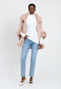 Kvinde iført lyserød kappe med pelskant, hvid turtleneck sweater, lyseblå jeans og hvide hælede mules, stående med den ene hånd i lommen.