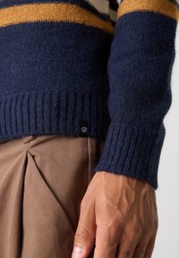Námornícky modrý vlnený sveter s oranžovými a krémovými pruhmi. Má rebrované manžety a spodný lem. Na dolnom leme je viditeľná malá čierna nášivka.
