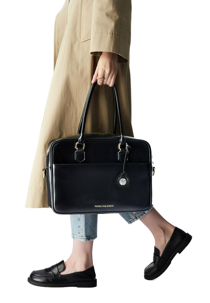 Portadocumenti in pelle nera con hardware dorato, forma rettangolare, tasca anteriore e un'etichetta con un orologio. Indossato con jeans blu chiaro e mocassini neri.
