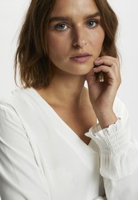 White, long-sleeve blouse with a V-neck. Features gathered cuffs and a soft, smooth texture. Close-up of a model's face and hand.