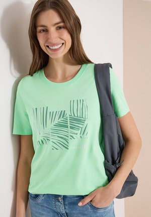 Mujer sonriente con una camiseta verde claro con diseño de hojas de palma, vaqueros azules y sujetando una chaqueta gris oscuro sobre su hombro.