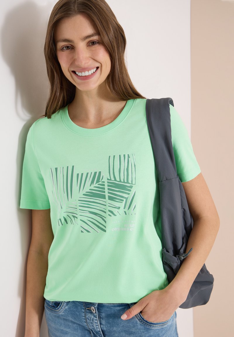 Femme souriante portant un t-shirt vert clair avec un motif de feuille de palmier, un jean bleu et tenant une veste gris foncé sur son épaule.