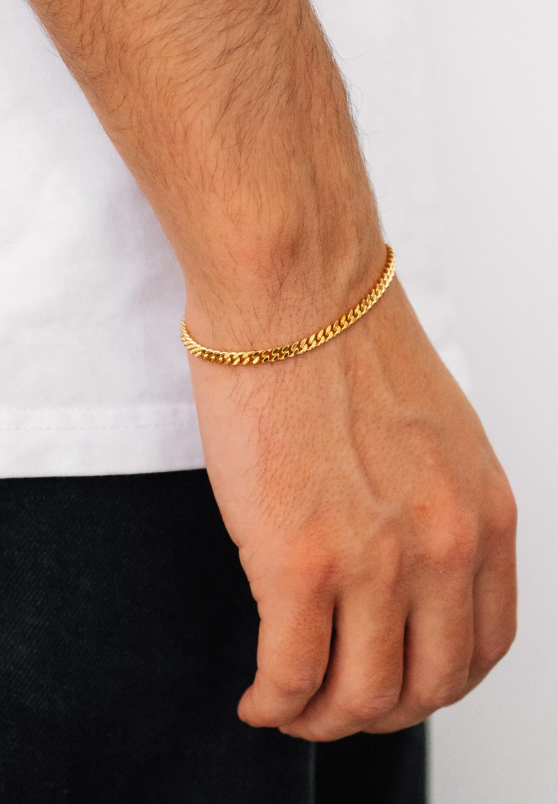 Gold chain bracelet on a wrist, featuring a twisted design with a smooth texture and shiny finish, against a plain background.