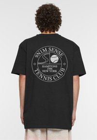 Melna T-krekls ar īsām piedurknēm, uz muguras ar apaļu balti izdrukātu logotipu. Uzraksts ietver "9N1M SENSE", "HAMPTONS" un "TENNIS CLUB".