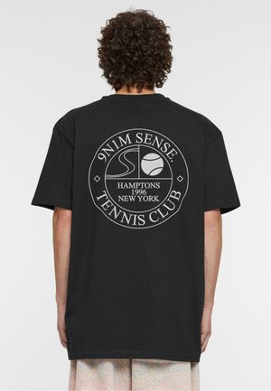 Sort t-shirt med korte ærmer, prydet med et cirkulært hvidt trykt logo på ryggen. Teksten inkluderer "9N1M SENSE", "HAMPTONS" og "TENNIS CLUB."