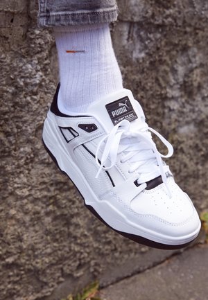 Zapatillas blancas Puma Slipstream con detalles negros, usadas con calcetines blancos y pantalones grises remangados, sobre un fondo de pared con textura rugosa.