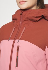 Röd och rosa syntetjacka med en dragkedjeförsedd bröstficka, justerbar huva och kontrasterande färgblock. Har texturerad struktur och dragsnören.