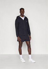 Άνδρας φορώντας μαύρο φερμουάρ μπουφάν, μαύρες βερμούδες, λευκό μπλουζάκι, λευκές κάλτσες και λευκά αθλητικά παπούτσια, που στέκεται μπροστά σε έναν απλό φωτεινό φόντο.