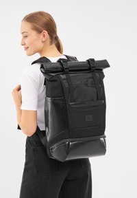 Fekete, felhajtható hátizsák fényes anyagból, struktúrált elülső panellel, állítható pántokkal és zipzáros alsó rekesszel.