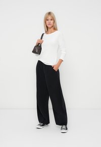 Top blanco de manga larga, pantalones anchos negros, sosteniendo un bolso oscuro, combinado con zapatillas en blanco y negro, frente a un fondo plano.