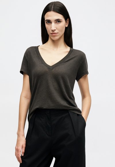 Femme aux longs cheveux bruns portant un t-shirt à manches courtes en V gris foncé, rentré dans un pantalon noir taille haute, debout avec une main dans la poche.