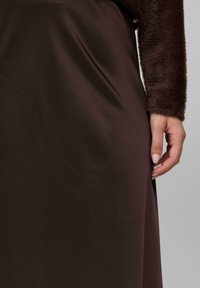Main dansante à côté d'une jupe en satin marron foncé, portant un haut à manches longues en peluche marron foncé sur fond neutre.
