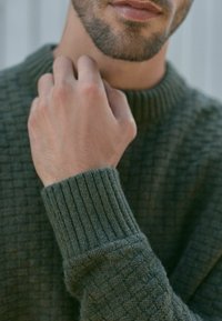 Sweter dzianinowy w ciemnozielonym kolorze z teksturowanym wzorem; zbliżenie na kołnierz i ściągacze, ukazujące miękki materiał przypominający wełnę. Ręka delikatnie dotyka szyi.