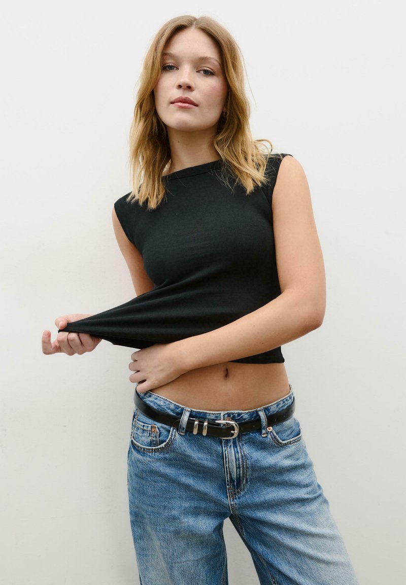 Jeune femme aux cheveux châtain clair portant un haut noir sans manches et un jean bleu, tirant son haut sur le côté sur un fond blanc uni.