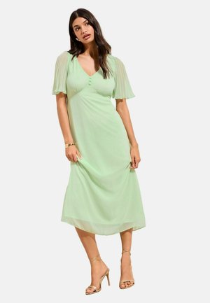 Femeie purtând o rochie midi verde deschis cu mâneci scurte transparente și sandale aurii cu toc înalt, stând în fața unui fundal alb.