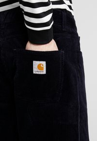 Pantaloni de catifea în nuanța navy, cu o patch pătrată ce prezintă un logo portocalie. Buzunar vizibil cu textură în relief și design drept.