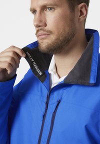 Blauwe Helly Hansen jas met een zwarte fleece-gevoerde kraag en rits. Heeft merknaam op de kraag en een gladde textuur.