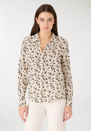 Blusa beige chiaro a maniche lunghe con stampa floreale nera, chiusura frontale con bottoni, colletto e tessuto morbido. Orlo frontale leggermente curvo.