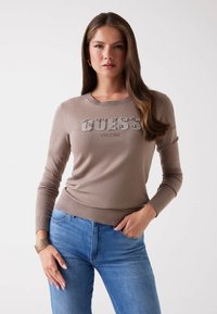 Pull en tricot marron avec logo "GUESS" orné de strass, col rond, manches longues et ourlet côtelé, porté avec un jean bleu.